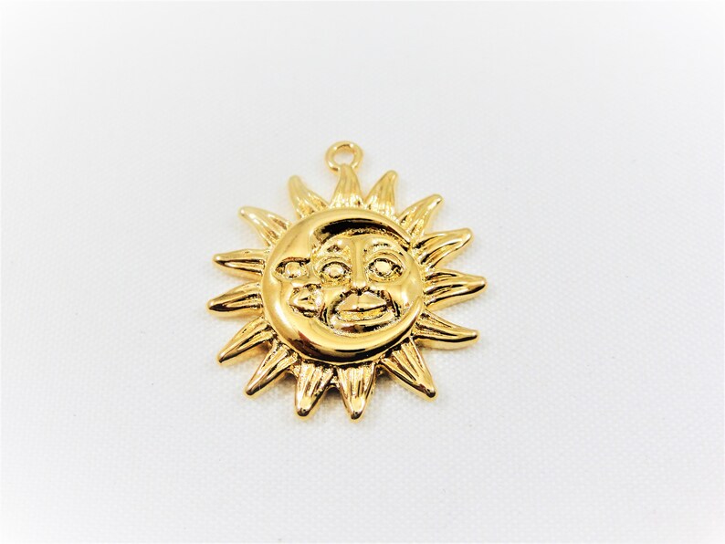 May include: Gold-toned pendant charm featuring a sun with a face and a crescent moon. The sun has a stylized face with a smile and eyes.