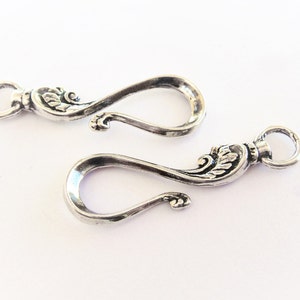 May include: Two silver-toned metal clasps with ornate floral designs. Each clasp has a loop on one end and a hook on the other.