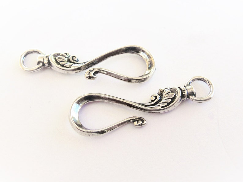 May include: Two silver-toned metal clasps with ornate floral designs. Each clasp has a loop on one end and a hook on the other.