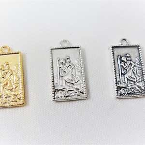 Pode incluir: Três pingentes retangulares com tema religioso. Um é dourado, os outros dois são prateados. Cada pingente apresenta um relevo detalhado de uma figura carregando outra pessoa, emoldurado por uma borda decorativa. Possuem uma pequena argola na parte superior para pendurar.