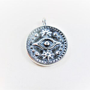 May include: Silver pendant with a round design featuring an eye with two stars on a white background.
