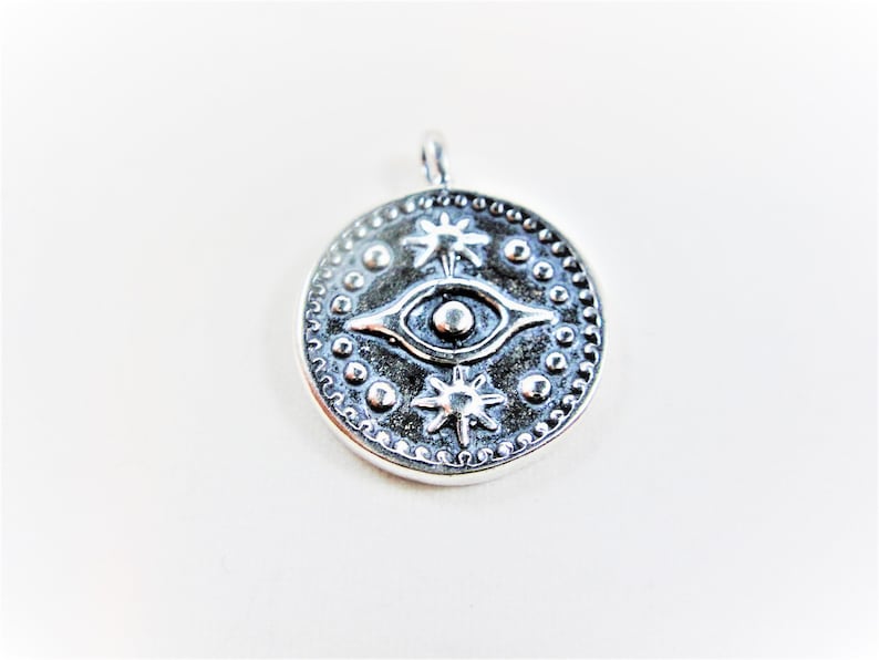 May include: Silver pendant with a round design featuring an eye with two stars on a white background.