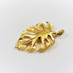 May include: A gold-colored metal pendant shaped like a monstera leaf. The pendant has a detailed design with cutouts and a textured surface.