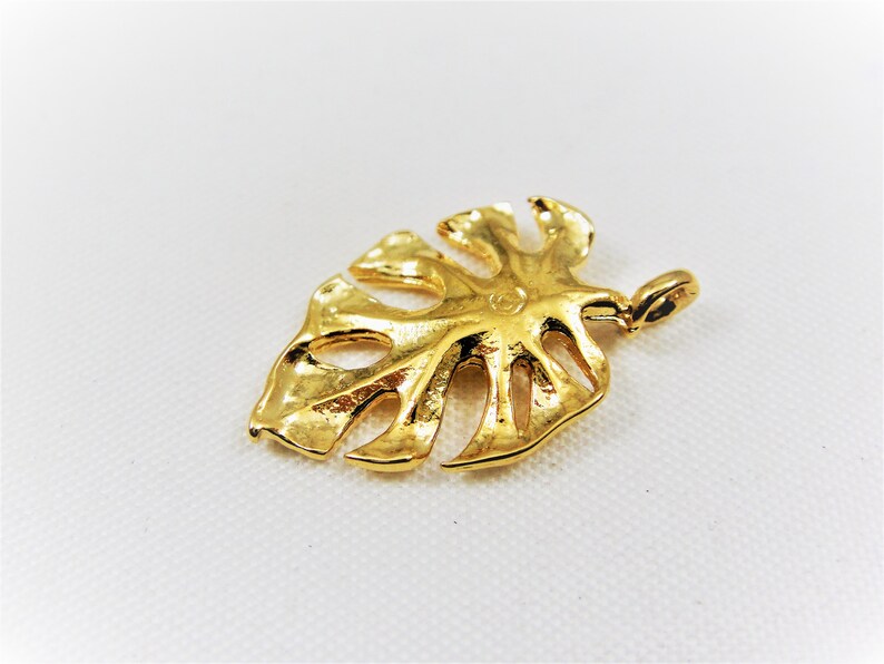 May include: A gold-colored metal pendant shaped like a monstera leaf. The pendant has a detailed design with cutouts and a textured surface.