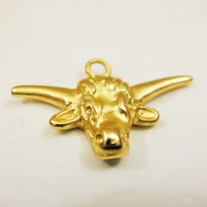 May include: A gold-colored metal charm in the shape of a bull's head. The charm has a loop at the top for attaching to a necklace or bracelet.