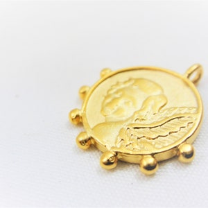 May include: Gold-toned pendant featuring a detailed relief of an angel with wings. The pendant is circular with a beaded edge.