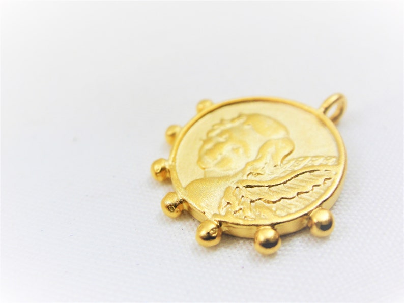 May include: Gold-toned pendant featuring a detailed relief of an angel with wings. The pendant is circular with a beaded edge.