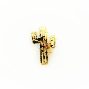 May include: A gold-colored cactus-shaped pendant with black and orange enamel detailing. The pendant has a small loop at the top for attaching to a chain or other jewelry. The cactus design is a popular southwestern motif.