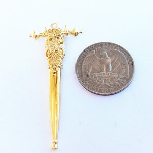 May include: A gold-colored metal sword charm with intricate details. The charm is about the size of a US quarter.