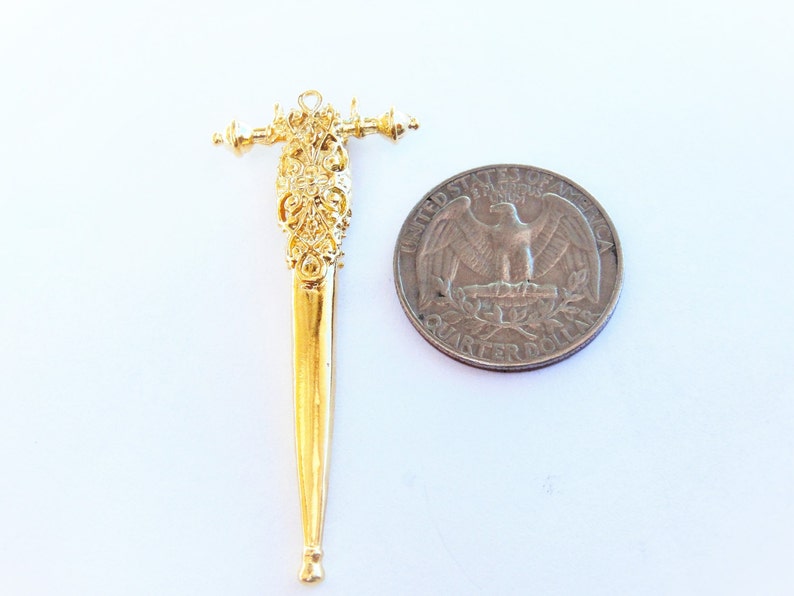 May include: A gold-colored metal sword charm with intricate details. The charm is about the size of a US quarter.