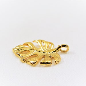 May include: A gold-colored metal charm in the shape of a monstera leaf with a loop at the top for attaching to a necklace or bracelet.