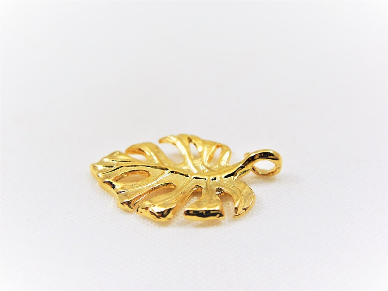 May include: A gold-colored metal charm in the shape of a monstera leaf with a loop at the top for attaching to a necklace or bracelet.