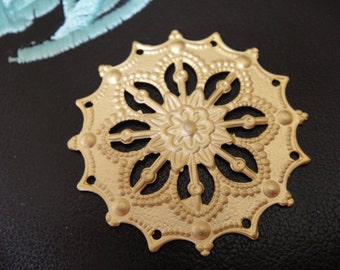 May include: A gold-coloured metal decorative embellishment with a circular, ornate design. The centre features a stylised flower with a raised, textured surface. The outer edges are scalloped and have a beaded border.
