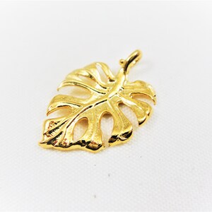 May include: A gold-colored metal pendant shaped like a monstera leaf. The leaf has a detailed design with cutouts and a textured surface.