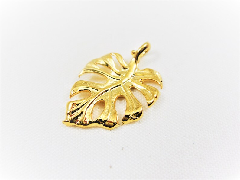May include: A gold-colored metal pendant shaped like a monstera leaf. The leaf has a detailed design with cutouts and a textured surface.