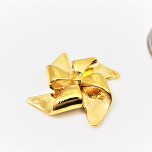 May include: Gold-toned metal charm shaped like a bow with four points, resembling a pinwheel. The charm has two holes for attaching to jewelry.
