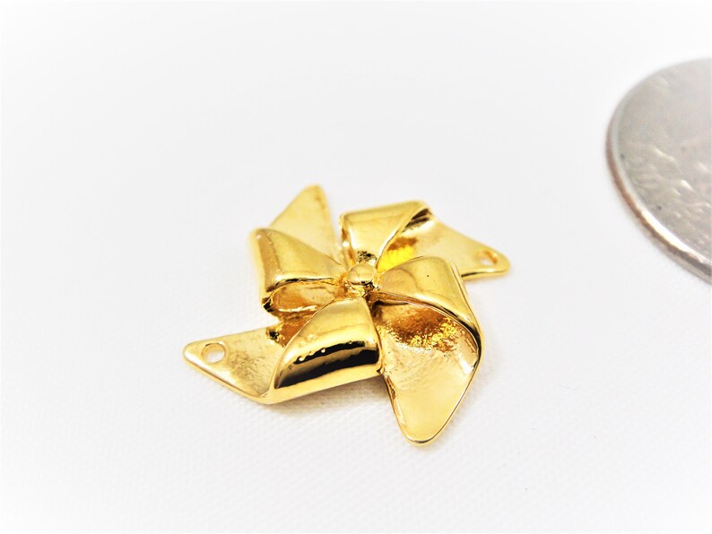 May include: Gold-toned metal charm shaped like a bow with four points, resembling a pinwheel. The charm has two holes for attaching to jewelry.