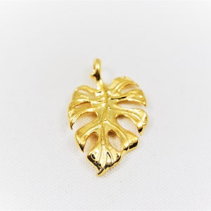 May include: A gold-colored pendant shaped like a monstera leaf. The pendant is detailed with cutouts and has a loop at the top for attaching to a chain.