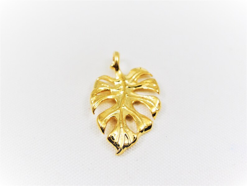 May include: A gold-colored pendant shaped like a monstera leaf. The pendant is detailed with cutouts and has a loop at the top for attaching to a chain.