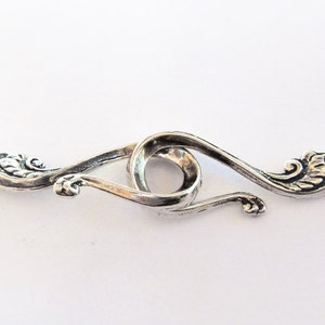 May include: Silver-toned metal clasp with a decorative design. The clasp has two loops that connect to form a circle in the center.