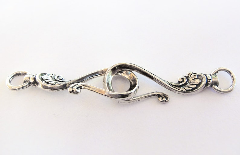 May include: Silver-toned metal clasp with a decorative design. The clasp has two loops that connect to form a circle in the center.