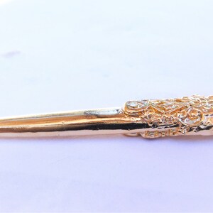 May include: A gold-colored metal kilt pin with a detailed, ornate design. The pin is shaped like a dagger with a pointed tip and a decorative handle.