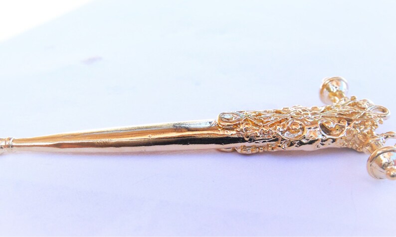 May include: A gold-colored metal kilt pin with a detailed, ornate design. The pin is shaped like a dagger with a pointed tip and a decorative handle.