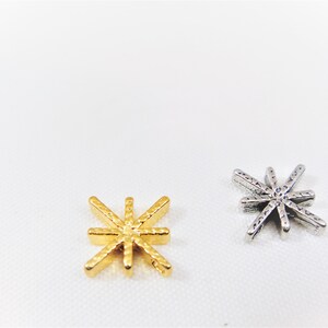 May include: Two star-shaped charms, one gold and one silver. Both charms have a textured surface and eight points.