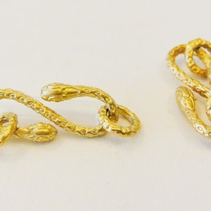 May include: Two gold-toned snake-shaped clasps for jewellery making. Each clasp is made of two intertwined snakes with detailed scales and heads.