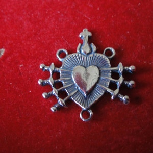 May include: Silver charm featuring a heart with seven swords piercing it, a cross on top, and two jump rings for attaching to a necklace or bracelet.