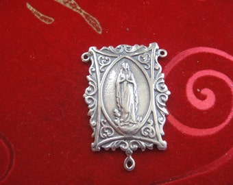 May include: Silver-toned pendant featuring a detailed image of the Virgin Mary in a rectangular frame with ornate scrollwork. The pendant has a loop at the bottom for attaching to a chain.