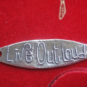 May include: A silver-colored metal charm with the words "Live Out Loud" engraved on it. The charm has two holes on either side.