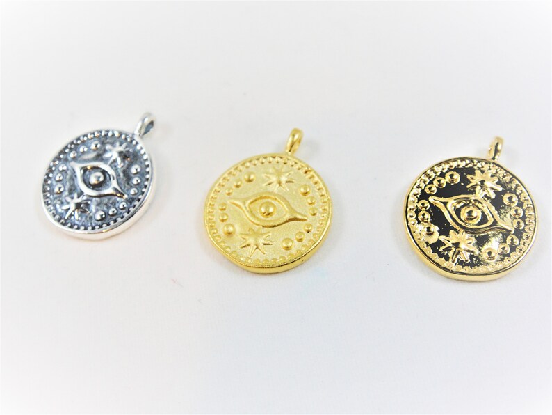 May include: Three round pendants with an eye design and star accents. The pendants are silver, gold, and gold with a black background.