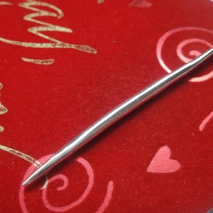 May include: A silver metal knitting needle on a red velvet background with a gold inscription and pink swirls.