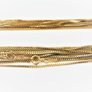 May include: A gold chain necklace with a box chain design. The chain is layered and folded in a pile.