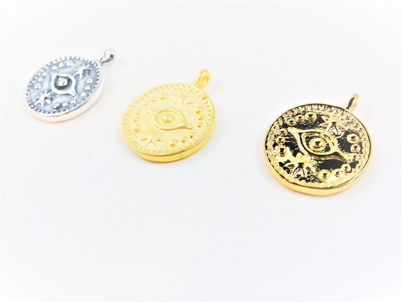 May include: Three gold and silver pendants with an evil eye design. The pendants are round and have a detailed design with a raised eye in the center.