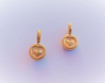 May include: Two gold-colored heart-shaped earrings. Each earring features a small, raised heart design within a circular frame, with a curved loop at the top for attachment. The earrings have a matte finish.