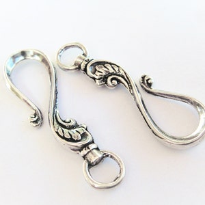 May include: Two silver-toned metal hooks with ornate floral designs. The hooks are designed to be used as clasps or closures for jewelry or other crafts.