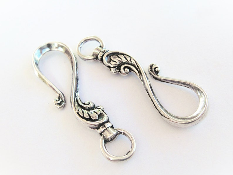 May include: Two silver-toned metal hooks with ornate floral designs. The hooks are designed to be used as clasps or closures for jewelry or other crafts.