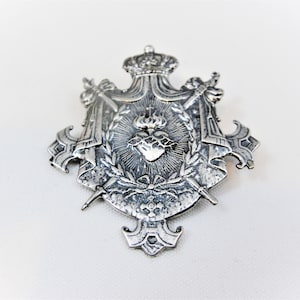 Puede incluir: Un emblema plateado detallado con una corona, espadas y un corazón con llamas. El emblema tiene forma de escudo con detalles intrincados, incluyendo una corona de laurel y pancartas decorativas. El diseño está sobre un fondo blanco.