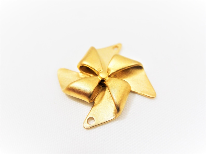May include: Gold-toned metal charm shaped like a bow with a pinwheel design. The charm has two holes for attaching to a necklace or bracelet.