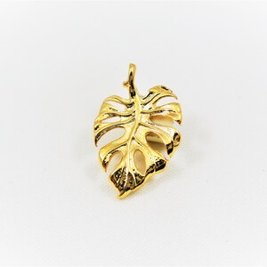 May include: A gold-colored pendant shaped like a monstera leaf. The pendant has a detailed design and is perfect for adding a touch of tropical style to any jewelry collection.