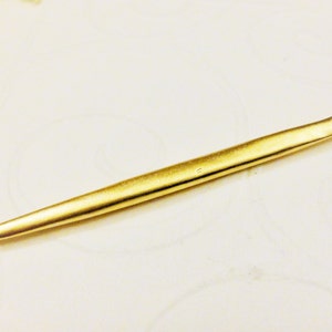 Könnte beinhalten: Eine goldfarbene Metallnadel mit abgerundeter Spitze und einer kleinen Schlaufe am anderen Ende.