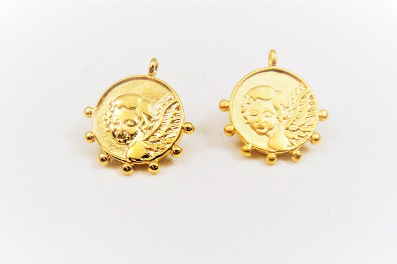 May include: Two gold-toned metal charms featuring a cherub with wings. Each charm has a circular design with a beaded border.