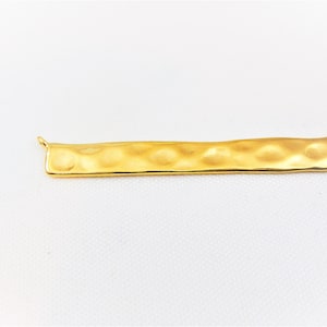 Puede incluir: Un colgante de barra de metal martillado de color dorado con dos anillos de salto para sujetarlo a un collar o pulsera.