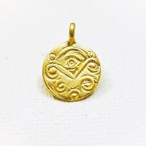 May include: A gold-colored pendant with an intricate, embossed design. The circular pendant features a central eye-like motif, surrounded by swirling patterns and a small loop for a chain. The pendant is likely made of metal.
