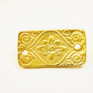 May include: A gold-coloured metal rectangular plaque with an ornate design. The plaque has two holes on the top edge.