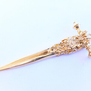 May include: A gold-colored, ornate, decorative dagger pendant with a detailed design. The pendant has a pointed tip and a decorative handle with a ball-shaped finial.