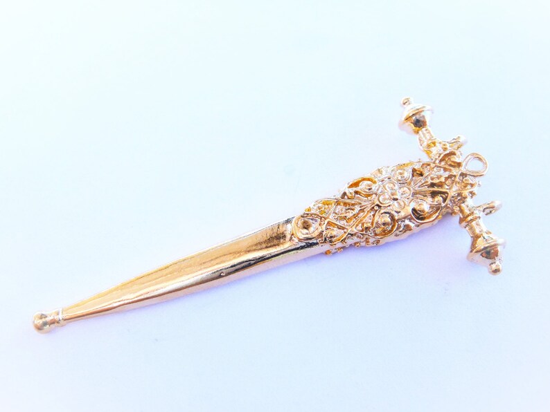 May include: A gold-colored, ornate, decorative dagger pendant with a detailed design. The pendant has a pointed tip and a decorative handle with a ball-shaped finial.