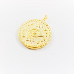 May include: Gold-toned pendant charm featuring a stylized eye with a small circle in the center, surrounded by stars and dots. The charm is round and has a textured surface.